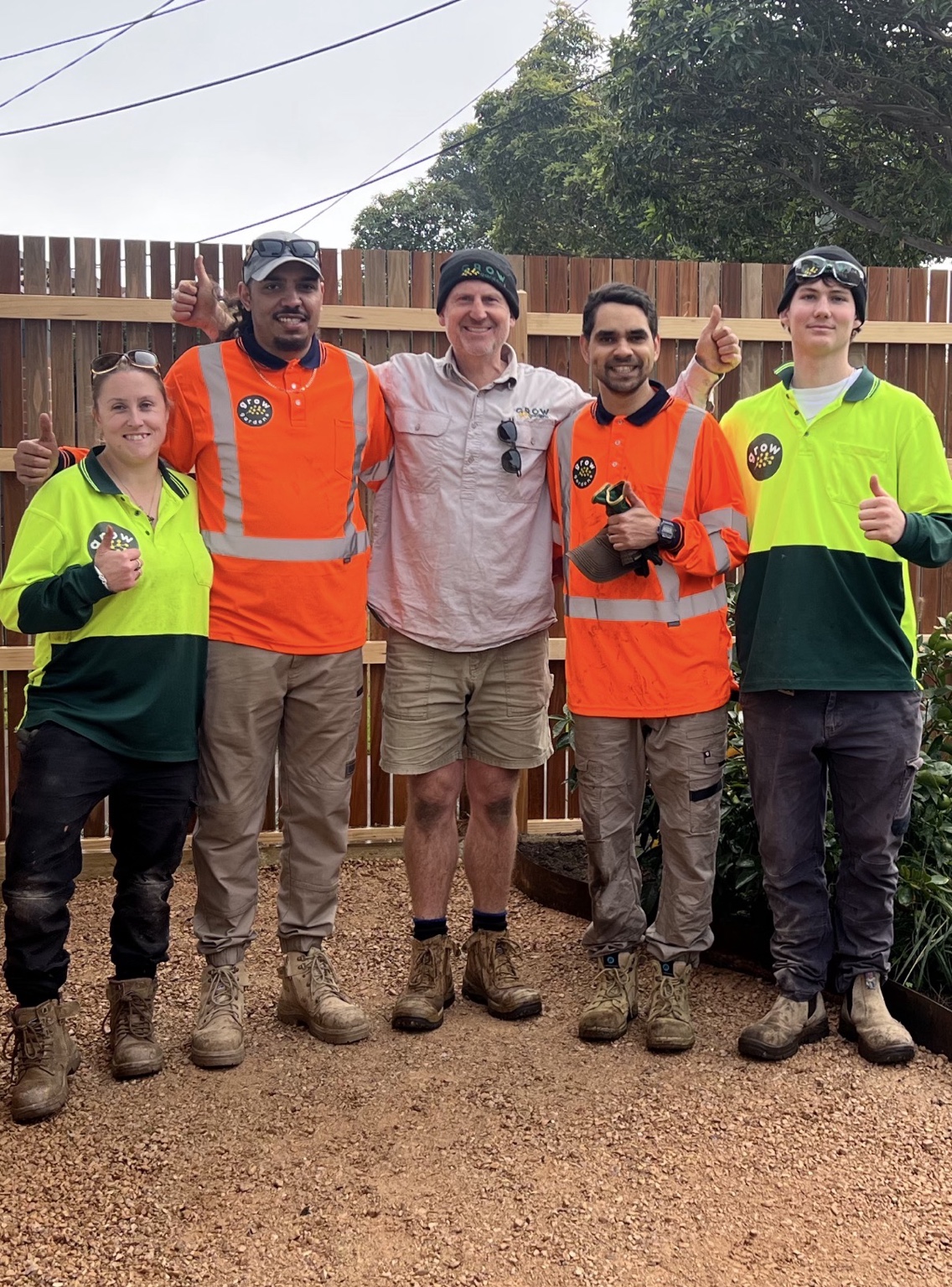 Five GROW Gardens team members smiling together in their work uniforms in a garden.